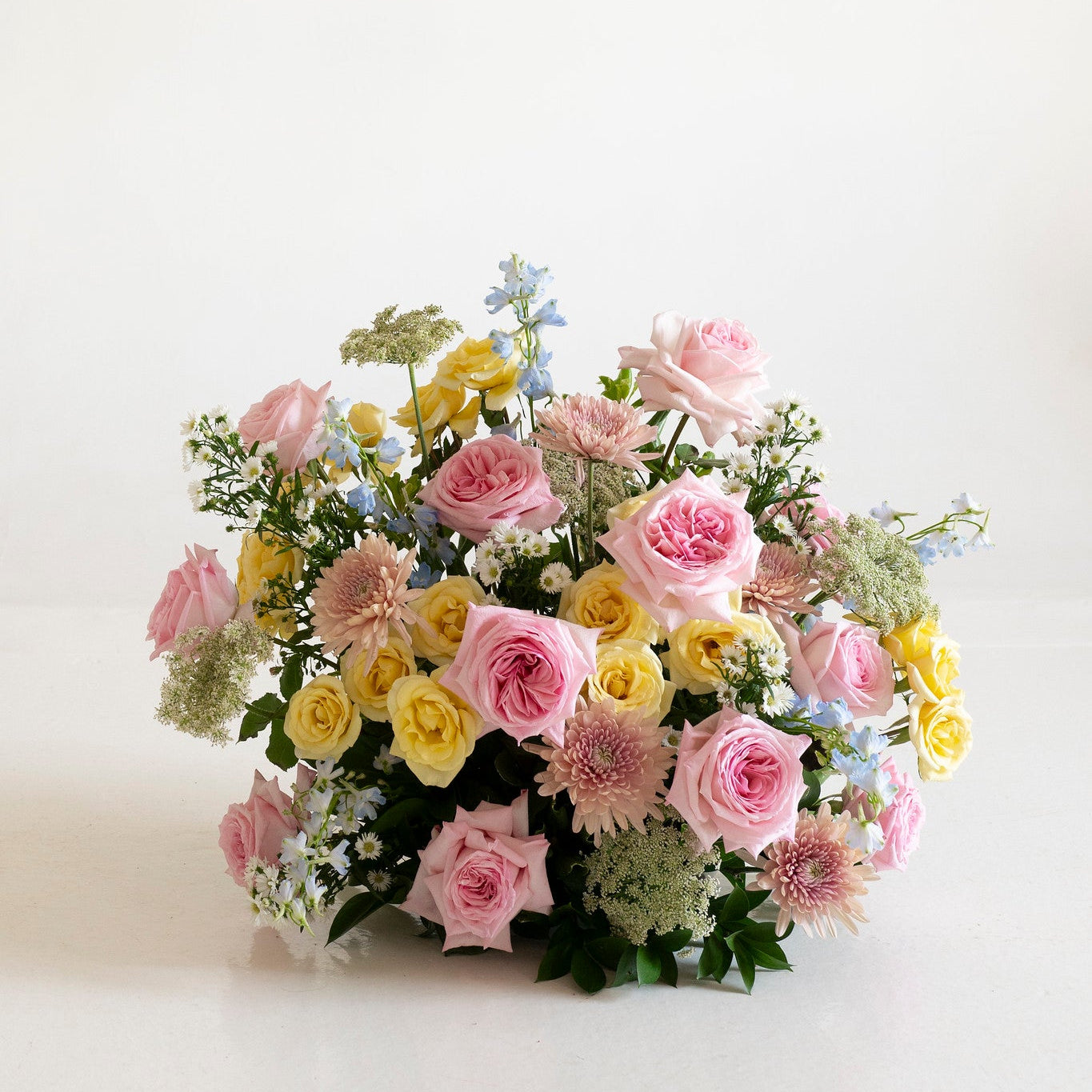 A pastel floral arrangement featuring pink and yellow roses, daisies, baby's breath, and green foliage, photographed on a clean white background.