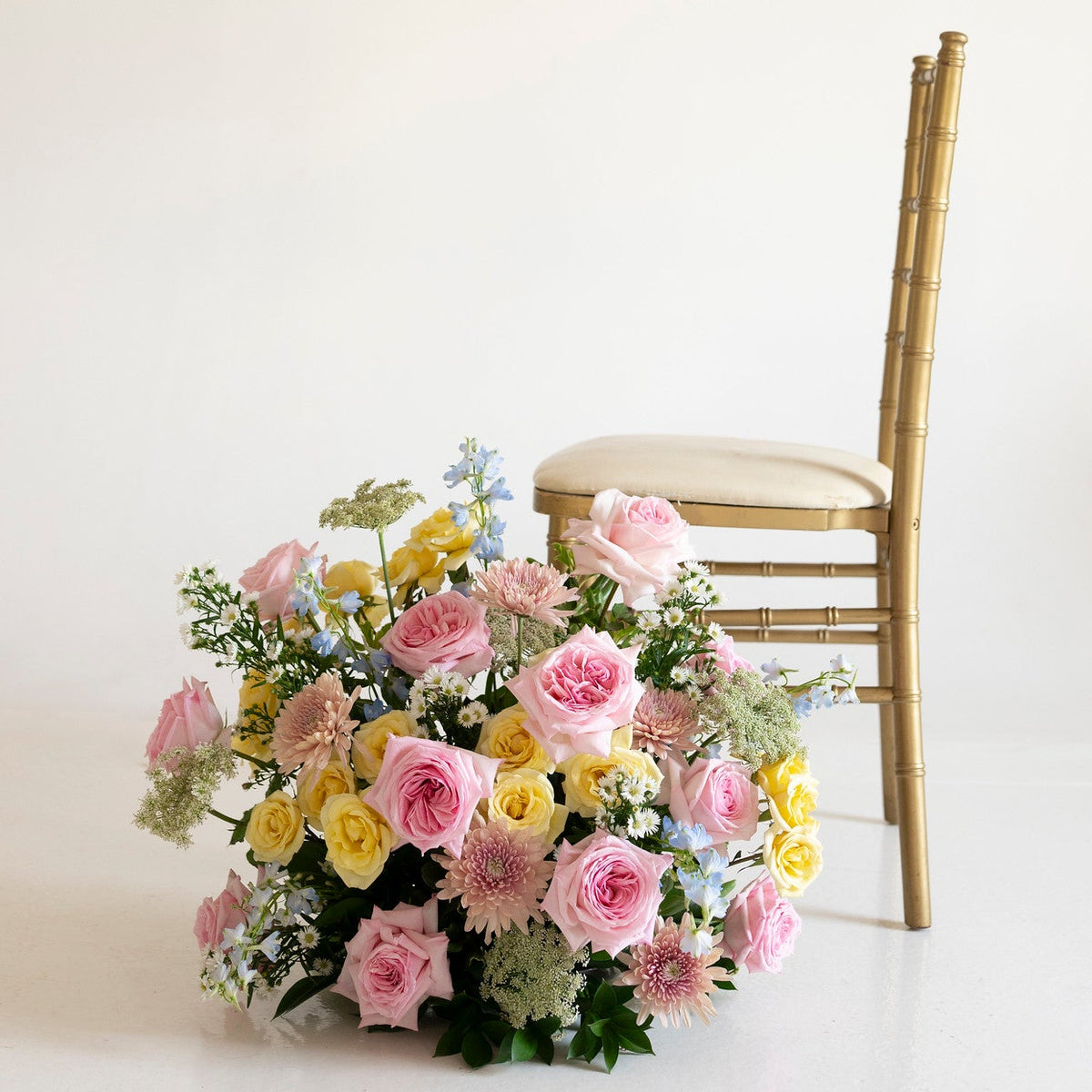 A pastel floral arrangement with pink, yellow, and light blue flowers sits on a white floor beside a gold bamboo-style chair against a plain off-white wall.