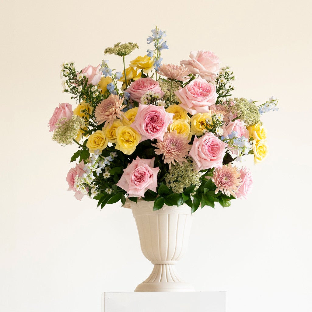A pastel flower arrangement in a white urn-shaped vase, displayed on a white pedestal against a clean off-white background. The bouquet features pink, yellow, and light green flowers with green foliage.