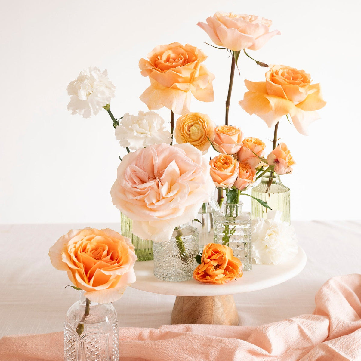 A styled floral arrangement of peach, orange, and white roses in clear glass vases and a white pedestal stand, set on a light fabric surface with a soft peach cloth in the foreground.