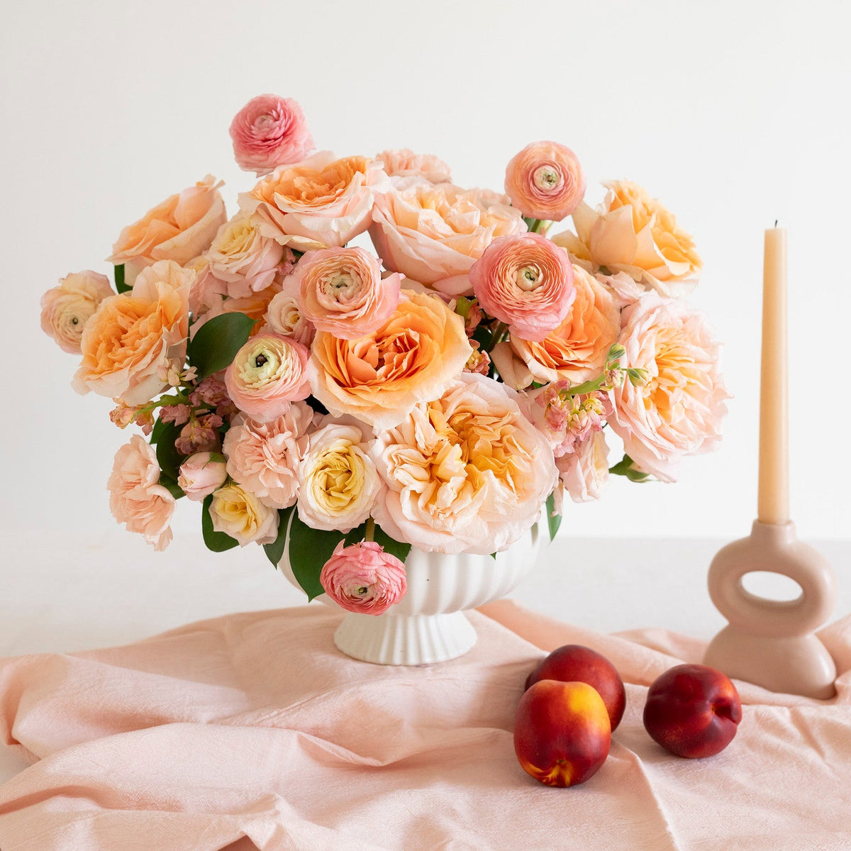 A bouquet of peach, pink, and cream roses in a white ribbed vase, styled on a draped blush pink fabric with two peaches and a beige candle holder against a white background.