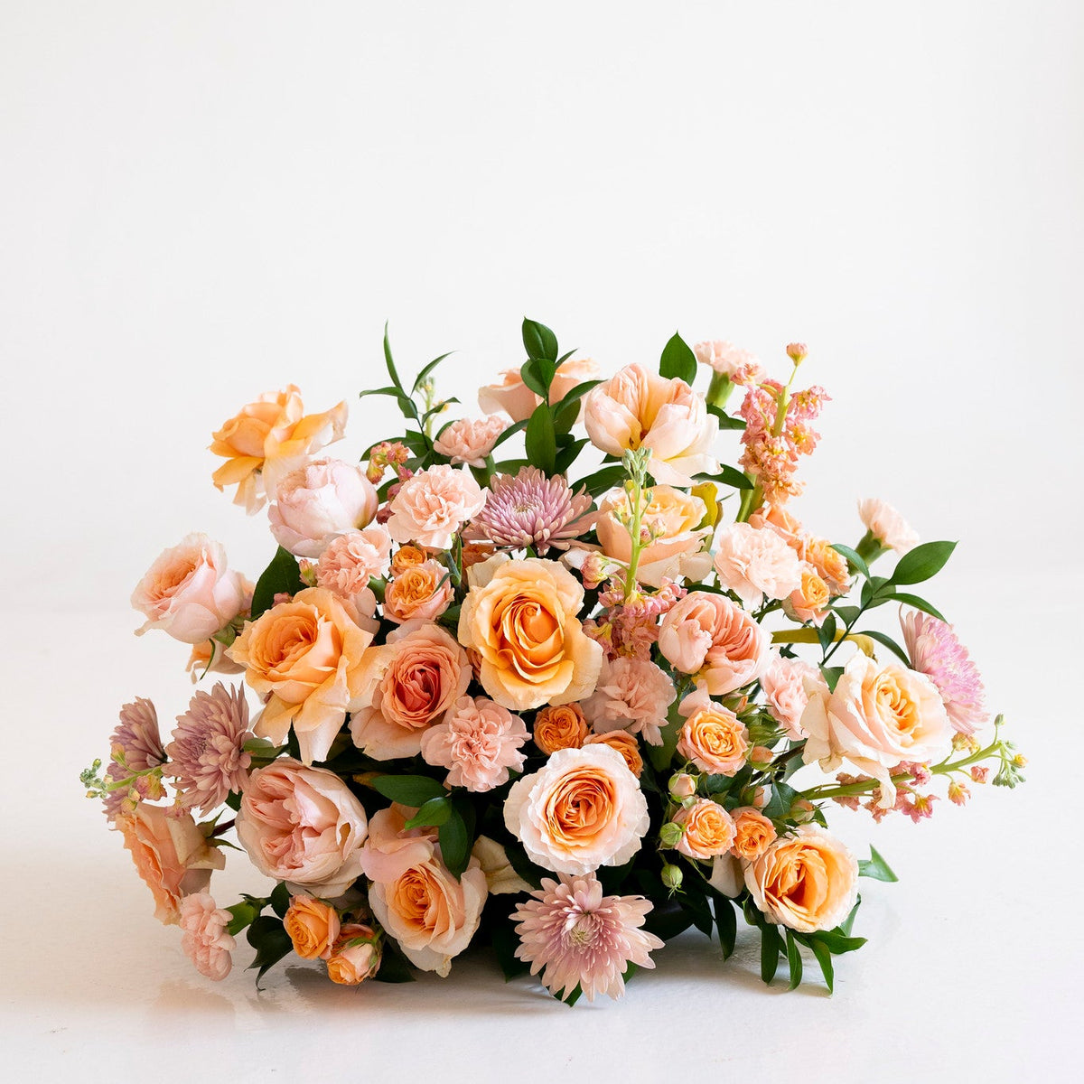 A lush floral arrangement featuring peach and orange roses, light pink chrysanthemums, and green foliage, set against a seamless white studio background.