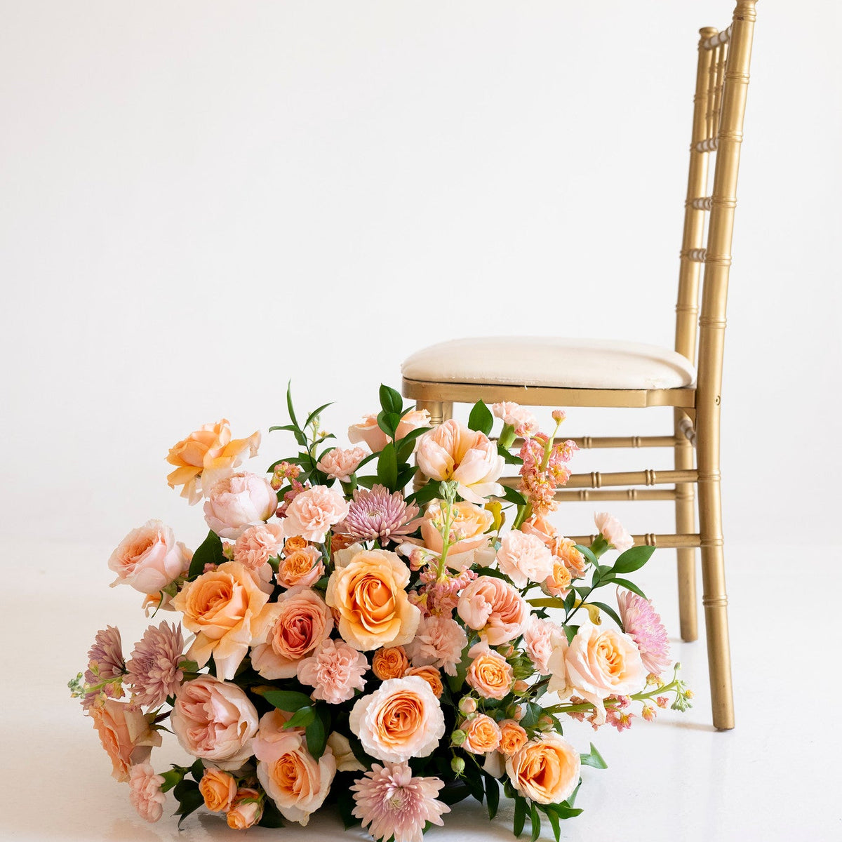 A large floral arrangement of peach, pink, and cream roses with green foliage sits on the floor beside a gold Chiavari chair against a clean white studio background.