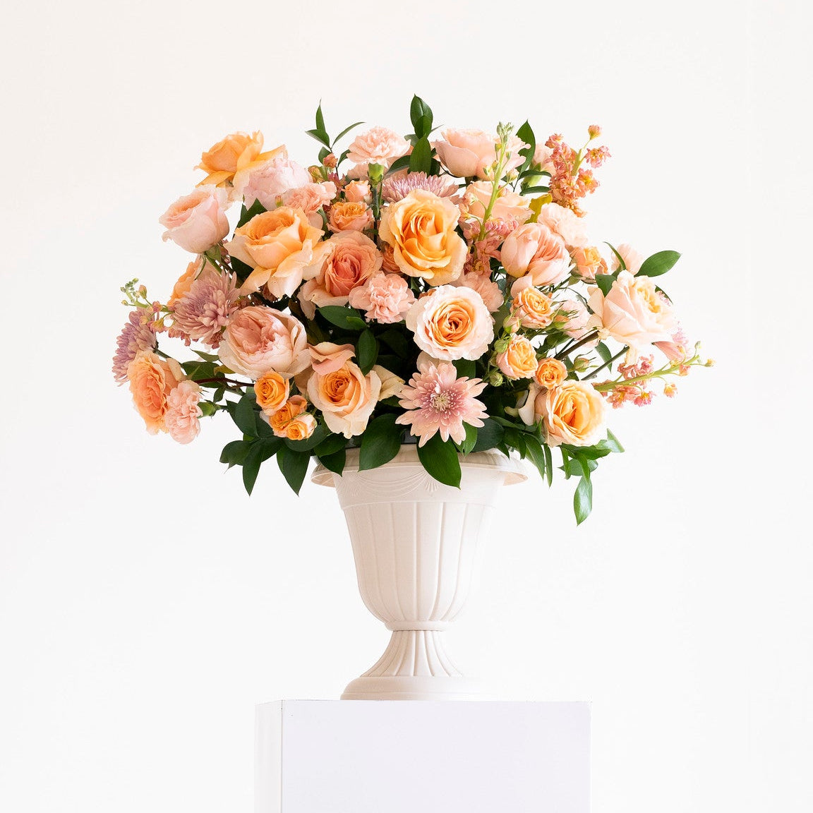 A bouquet of peach, pink, and cream roses with green foliage arranged in a white urn-shaped vase, displayed on a white pedestal against a white background.