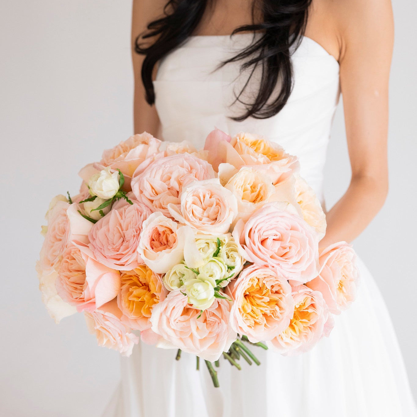 A woman in a white strapless wedding dress holding a large bouquet of soft peach, blush, and white roses against a plain light background.