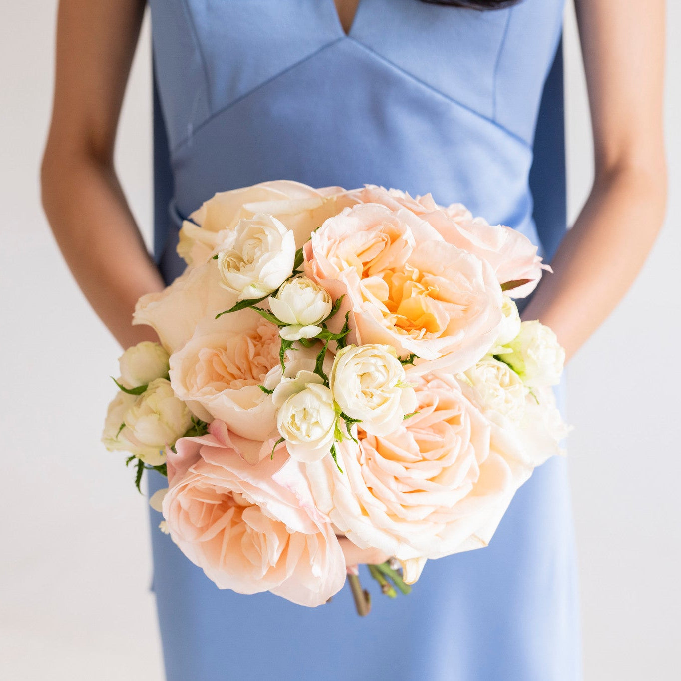 A bouquet of blush peach and cream roses held by a person wearing a light blue dress against a plain light background.