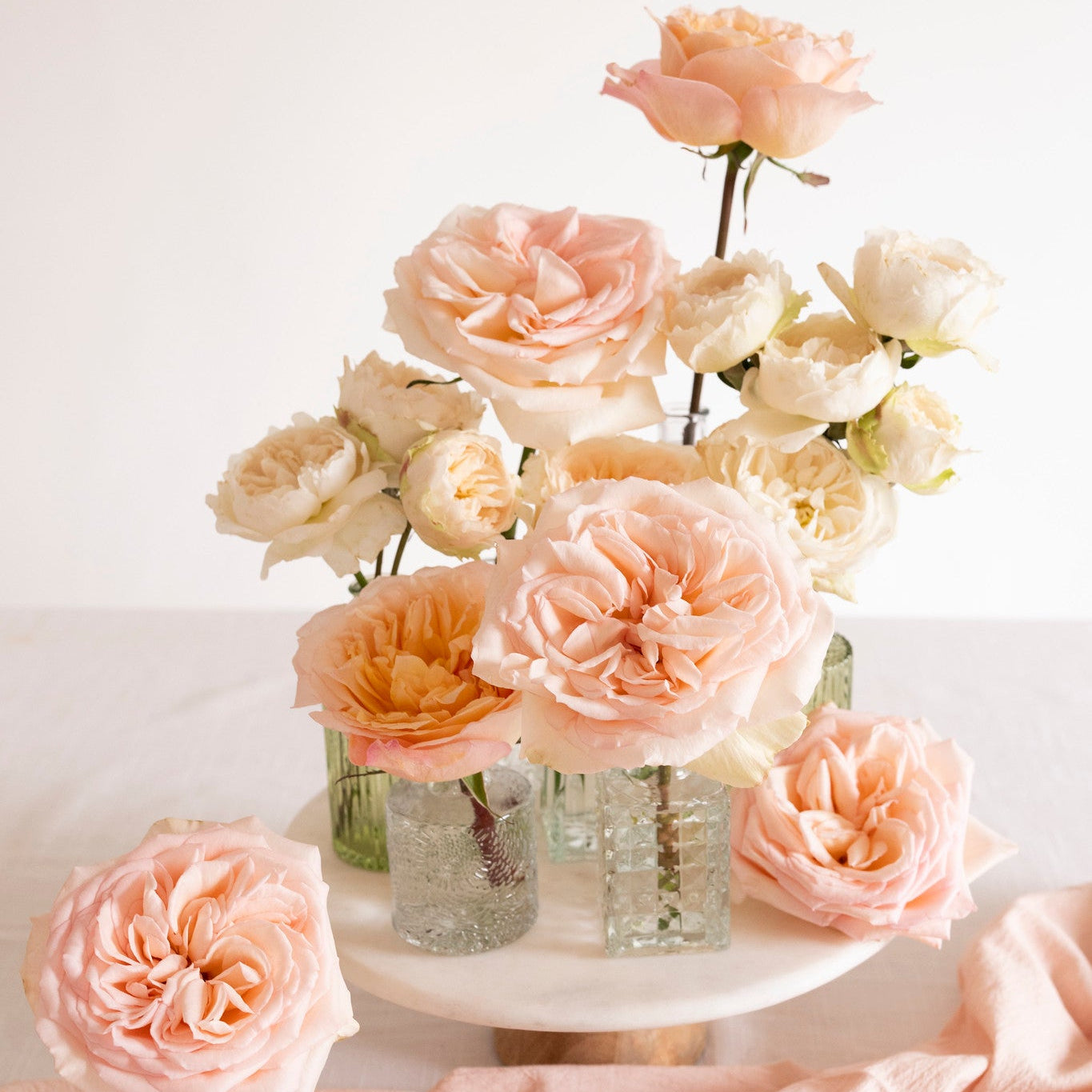 A styled floral arrangement of blush pink and cream roses in clear textured glass vases, displayed on a white pedestal with draped blush fabric in the foreground.