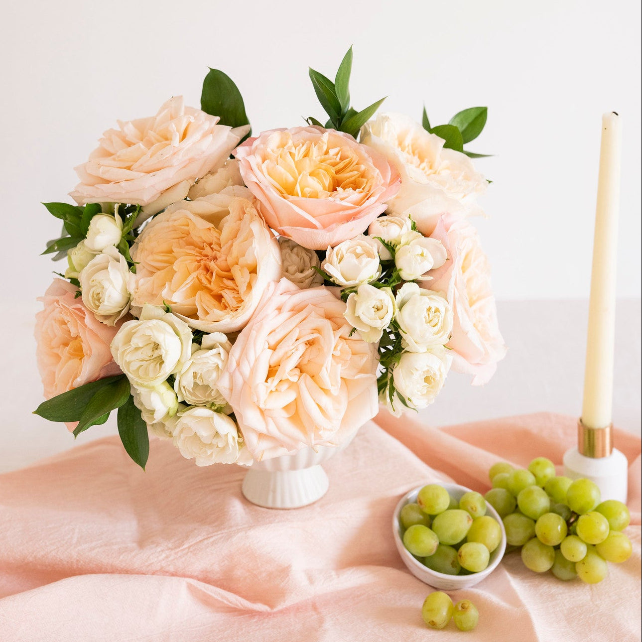 A bouquet of peach and cream roses with green leaves arranged in a white pedestal vase on a draped blush pink fabric, with green grapes and a white taper candle beside it against a soft white background.