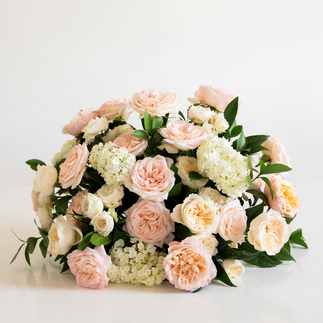 A round floral arrangement featuring blush pink and cream roses, white hydrangeas, and green foliage, photographed on a clean white surface against a white background.