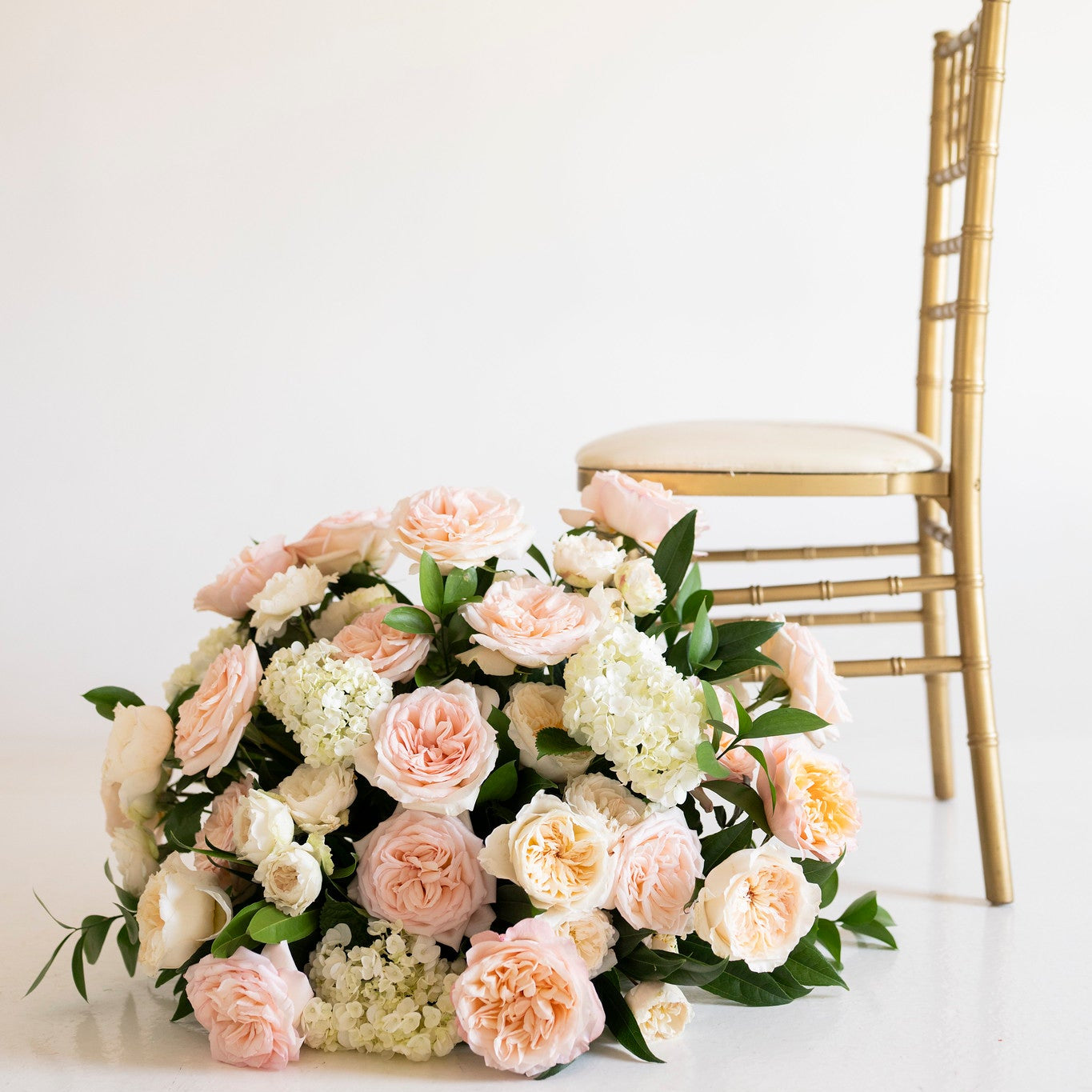 A large floral arrangement of blush pink, peach, and white roses with green foliage sits on a white floor beside a gold-framed chair against a plain off-white wall.