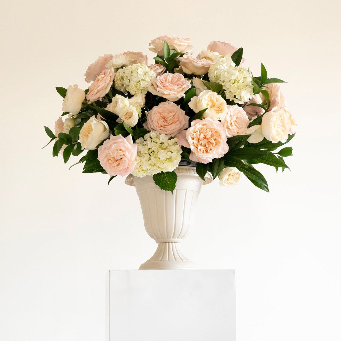 A bouquet of pale blush pink and cream roses with green foliage arranged in a white pedestal vase, displayed on a white rectangular plinth against a clean white background.