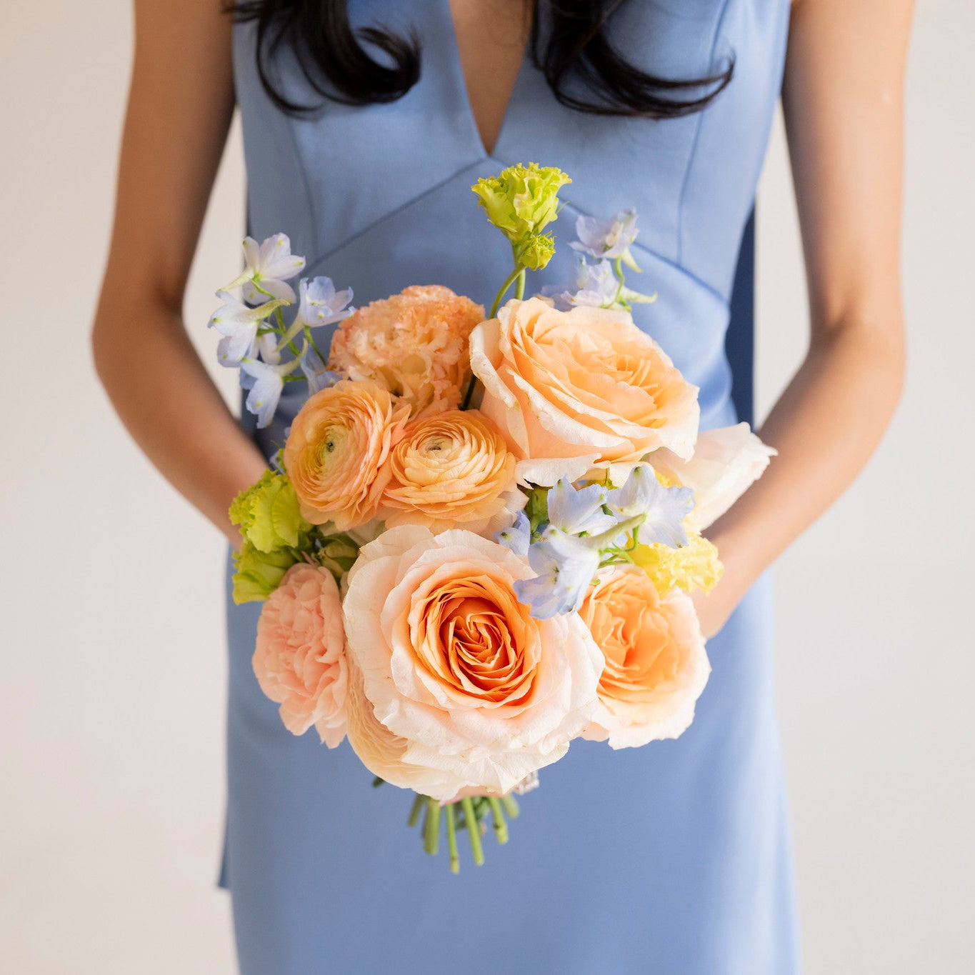 A woman in a light blue sleeveless dress holds a bouquet of peach and cream roses with light blue flowers and green stems against a plain light background.
