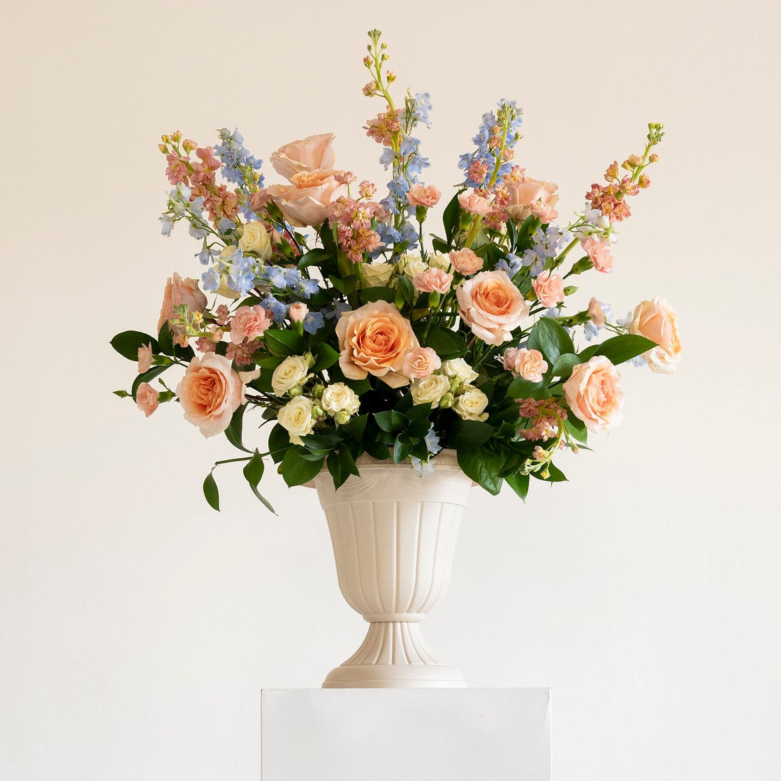 A floral arrangement in a white urn-shaped vase displayed on a white pedestal against a warm off-white background.