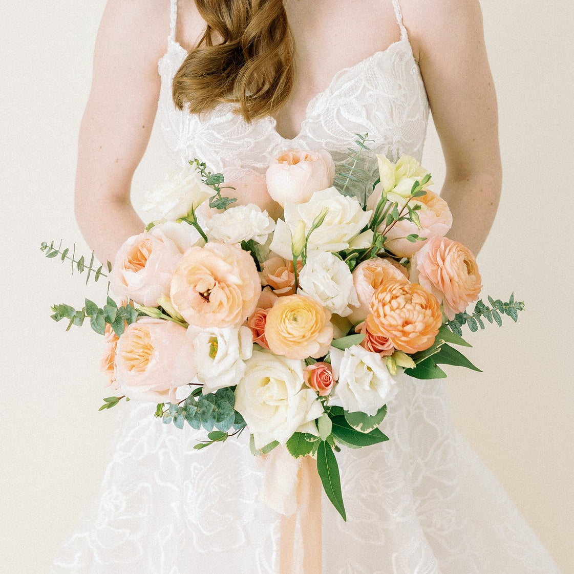 A woman in a white lace wedding dress holding a pastel peach and cream floral bouquet, photographed against a soft off-white studio background.