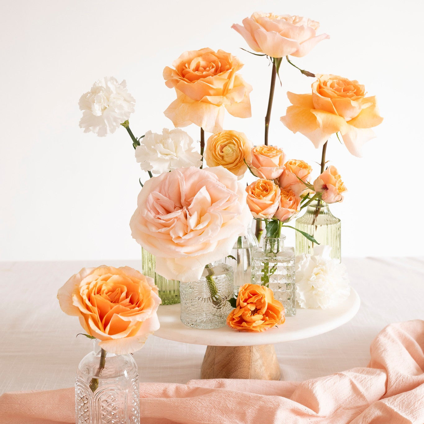 A styled floral arrangement of peach, orange, and white roses in clear glass vases and a white pedestal stand, set on a light fabric surface with a soft peach cloth in the foreground.