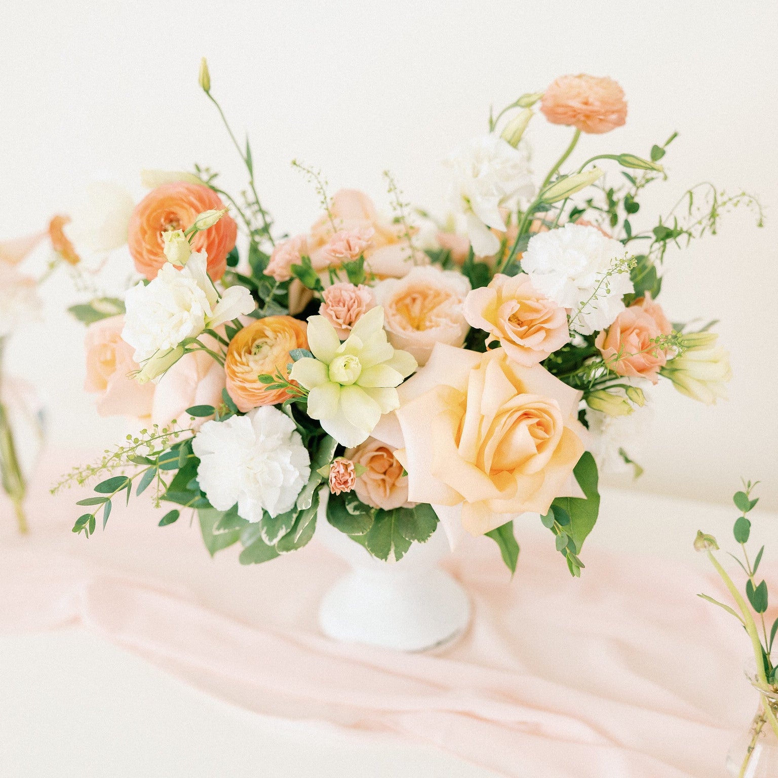 A soft floral arrangement in a white vase on a pale pink draped table runner, with peach, cream, and white flowers mixed with green foliage. A white taper candle stands to the right against a warm off-white background.