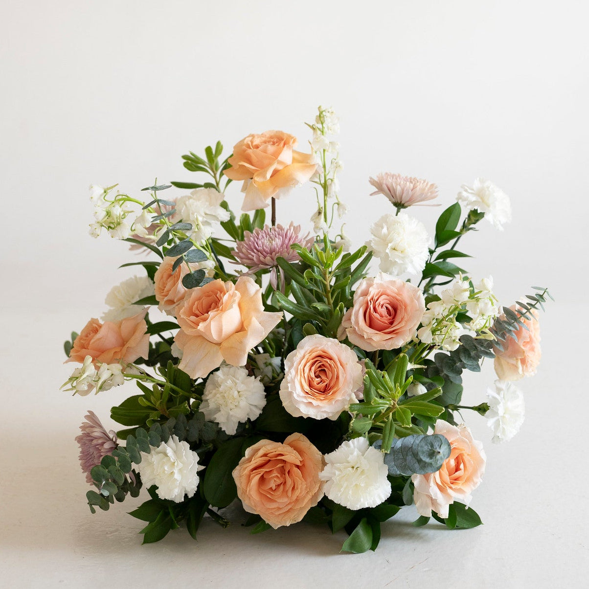 A round floral arrangement featuring peach and white roses, white carnations, and green foliage, set against a clean off-white background.