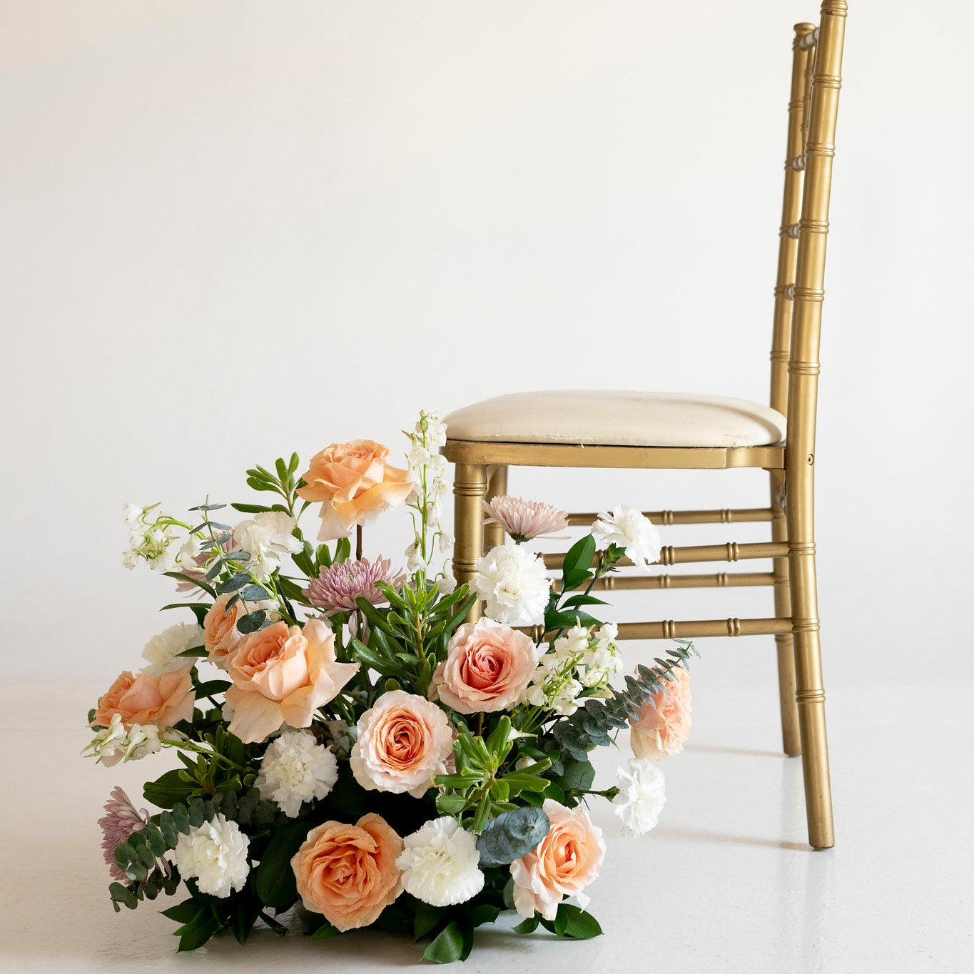 A gold bamboo-style chair with a cream upholstered seat, styled beside a large floral arrangement of peach, white, and blush roses with green foliage on a clean white studio background.