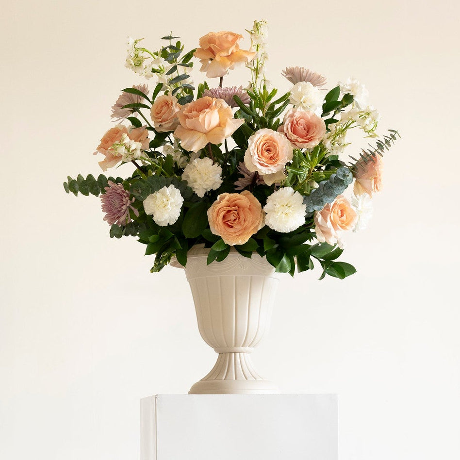 A floral arrangement in a white urn-style vase displayed on a white pedestal against a warm off-white background.