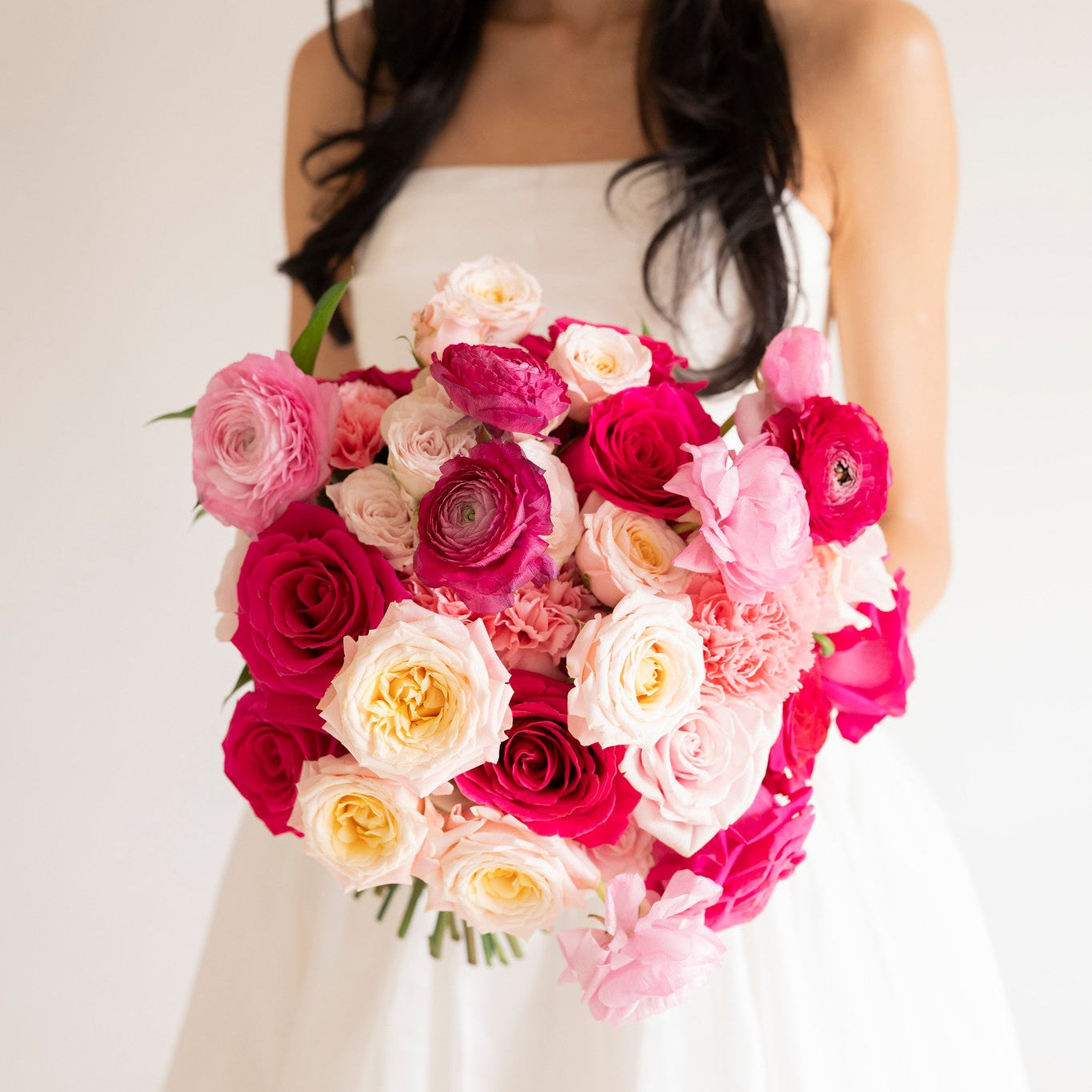 A woman in a white strapless dress holding a large bouquet of pink, magenta, and cream roses against a plain light background.