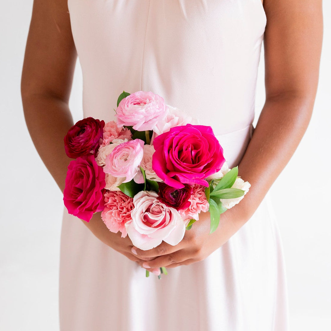 A woman wearing a pale blush pink halter-neck dress holds a vibrant bouquet of pink and red roses against a clean white background.