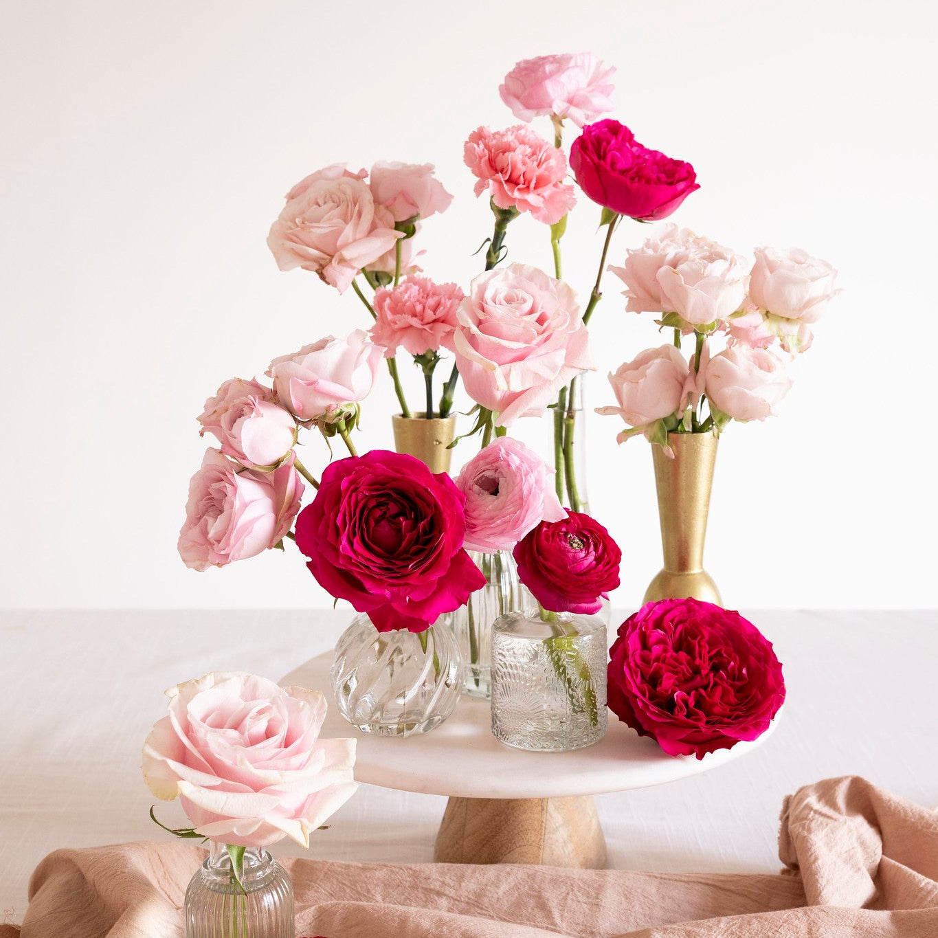 A styled floral arrangement of pink and deep red roses in clear glass vases and a gold vase, displayed on a white pedestal with a draped blush fabric underneath against a white background.