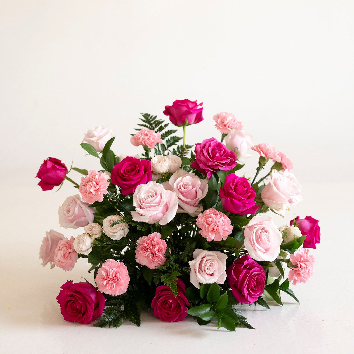 A mixed floral arrangement featuring pink and magenta roses, light pink carnations, and green foliage, displayed on a clean white surface against a white background.
