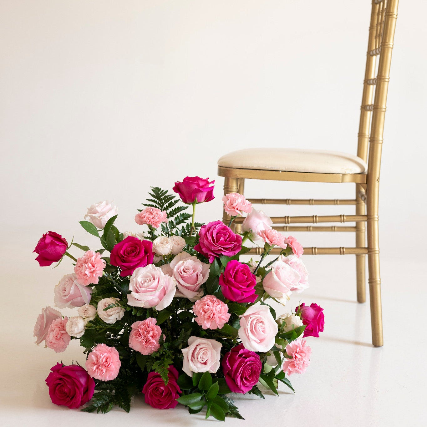 A large floral arrangement of pink, magenta, and white roses with green foliage sits on a white floor beside a gold Chiavari chair against a plain off-white background.