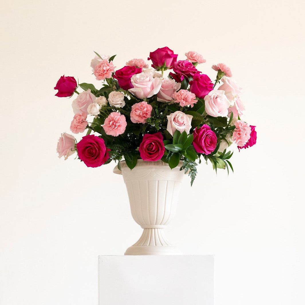 A bouquet of pink and magenta roses with green foliage arranged in a white urn-shaped vase, displayed on a white rectangular pedestal against a clean white background.
