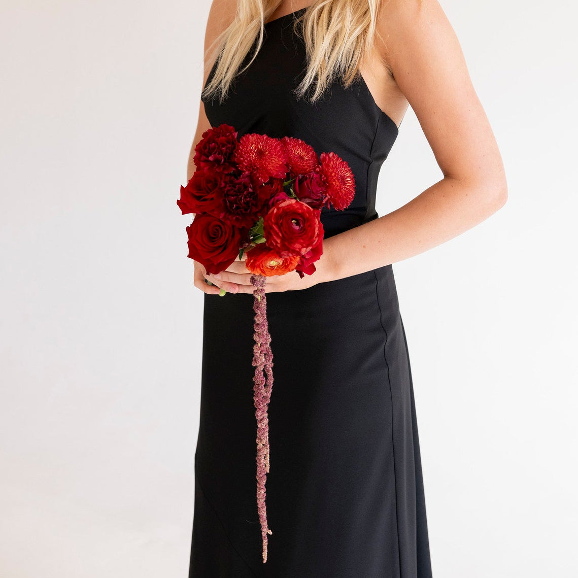 A blonde woman wearing a sleeveless black dress holds a bouquet of deep red flowers against a plain light background.