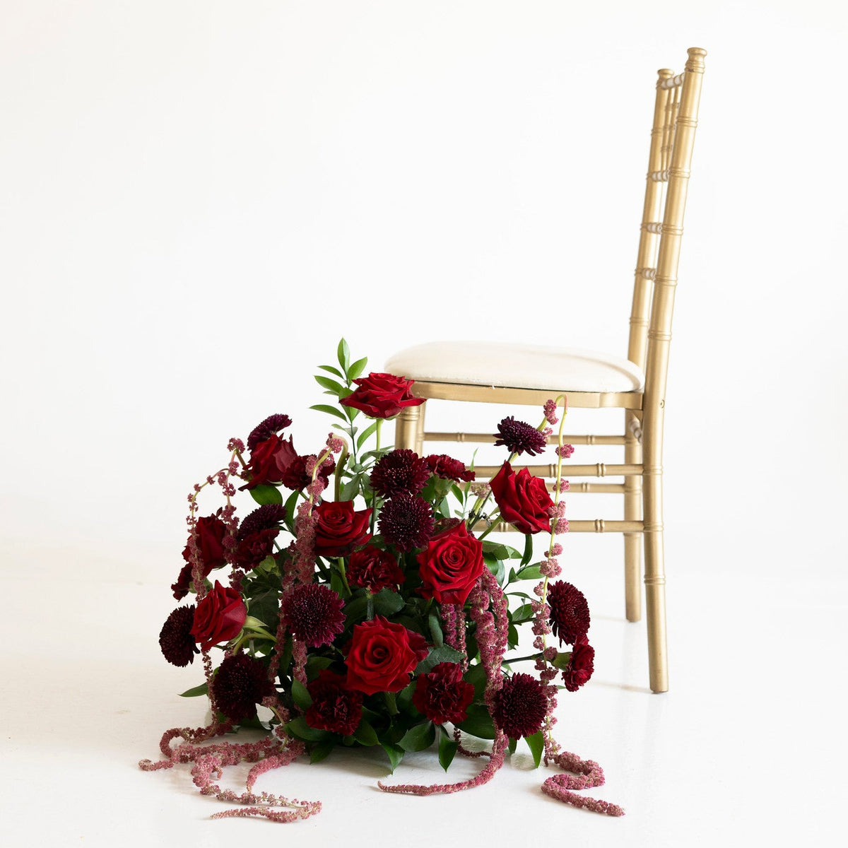 A large floral arrangement of deep red and burgundy roses with green foliage sits on a white floor beside a gold Chiavari chair against a seamless white background.