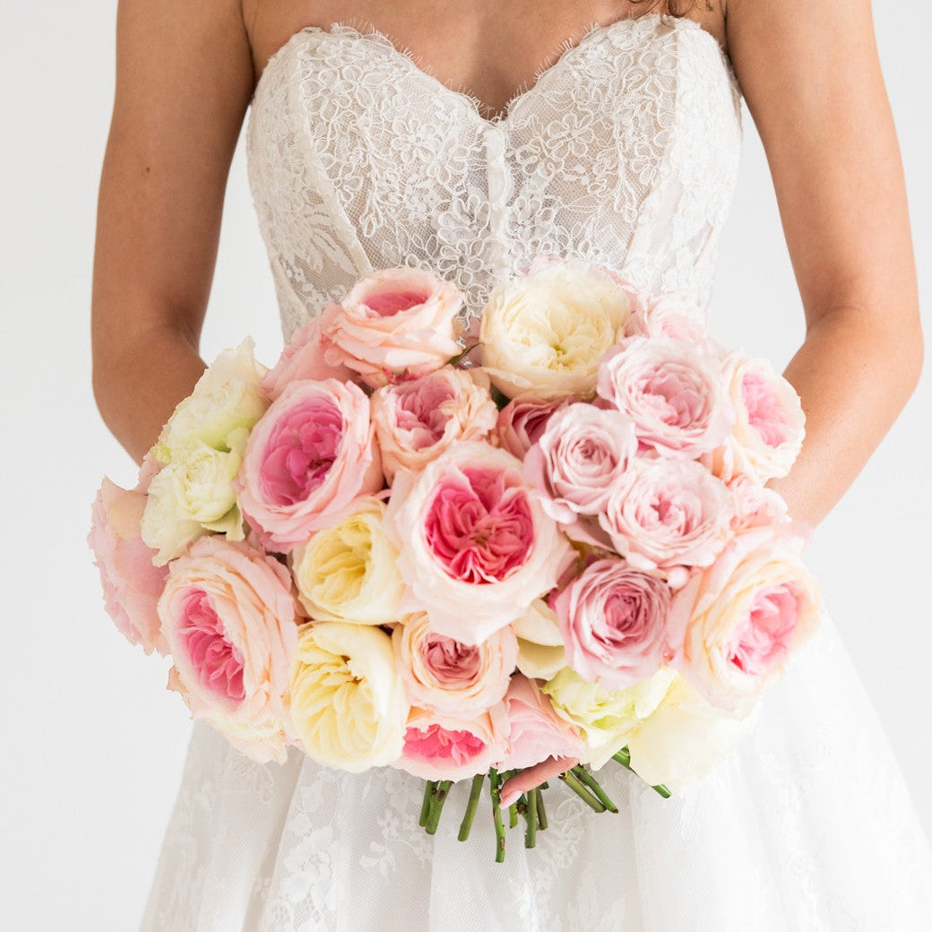 A bride in a strapless white lace wedding dress holding a large bouquet of pink, blush, and cream roses against a plain white background.