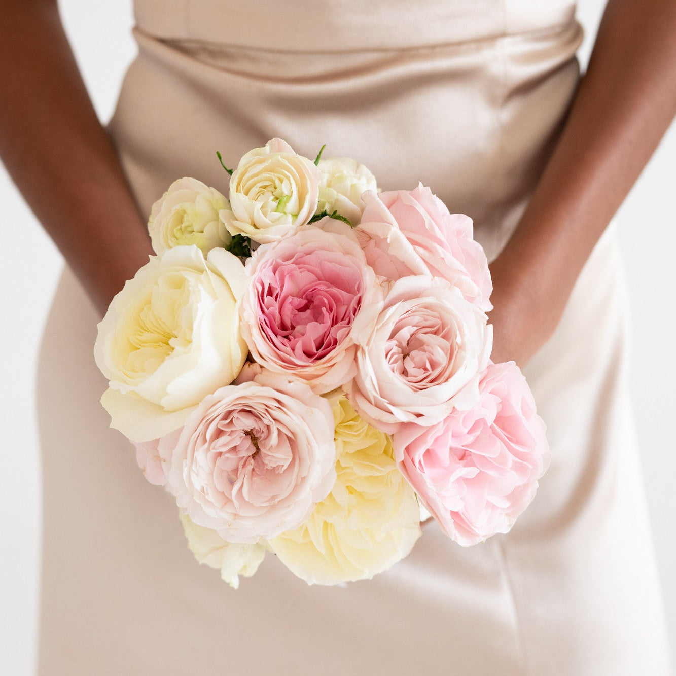 A bouquet of soft pink and cream peonies held by a person wearing a champagne satin dress against a clean white background.