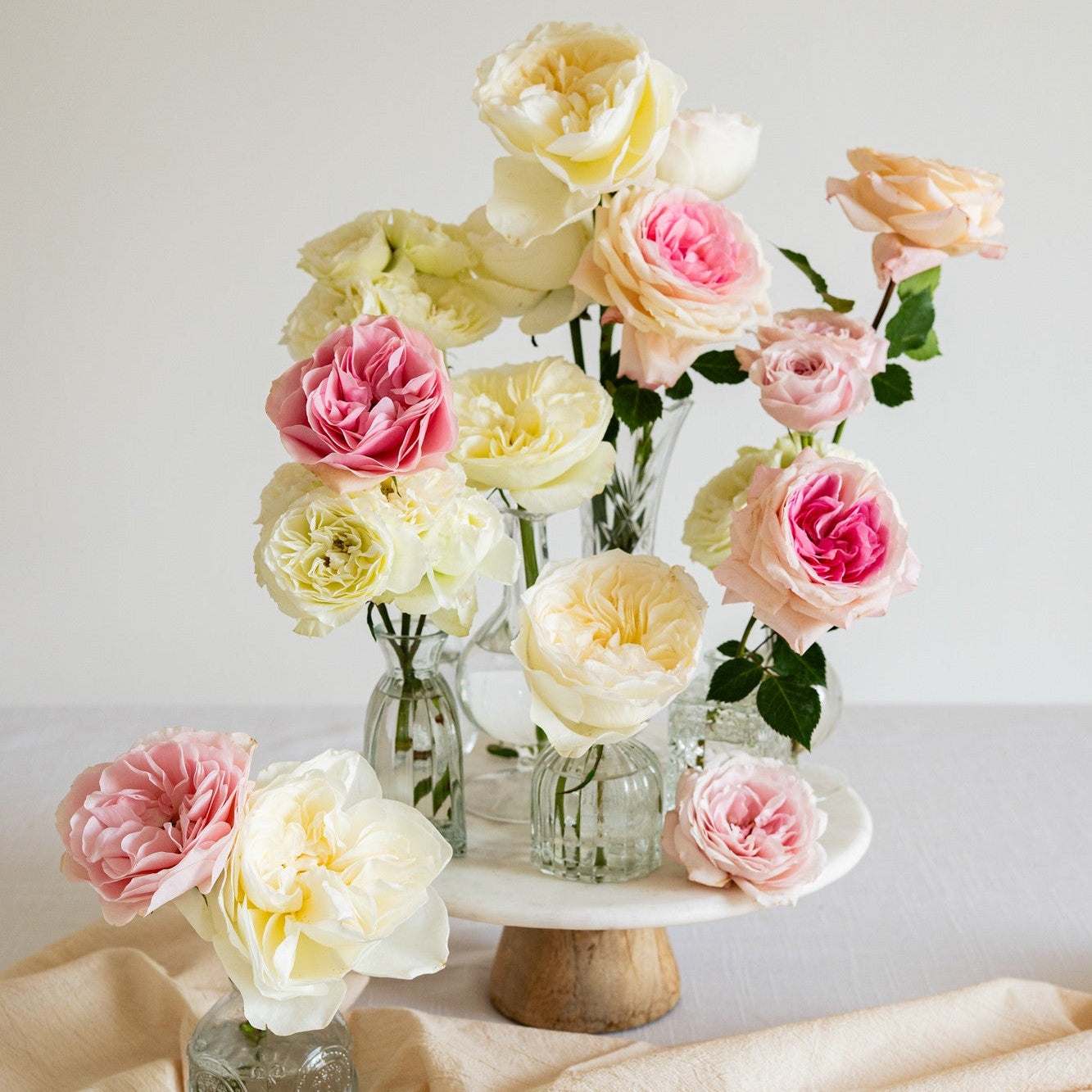 A floral arrangement of pale yellow and pink roses in clear glass vases, displayed on a white pedestal stand with a beige fabric drape against a soft neutral background.