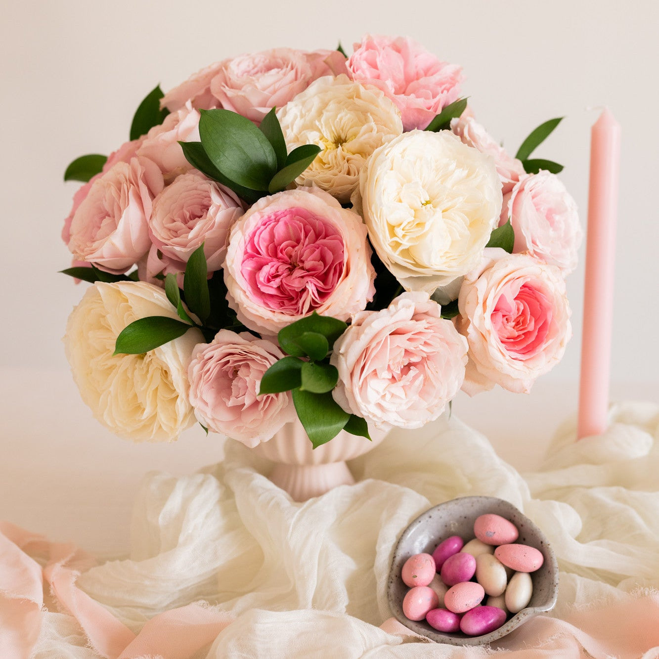 A bouquet of pink and cream roses with green leaves arranged in a vase, set on draped pale pink fabric with a small bowl of pink candies and a pink taper candle nearby against a soft cream background.