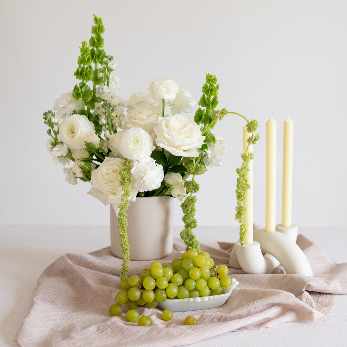 A styled floral arrangement in a white vase with white roses and green stems, set on a pale pink cloth beside white candlesticks and a small dish of green grapes on a white surface against a light neutral background.