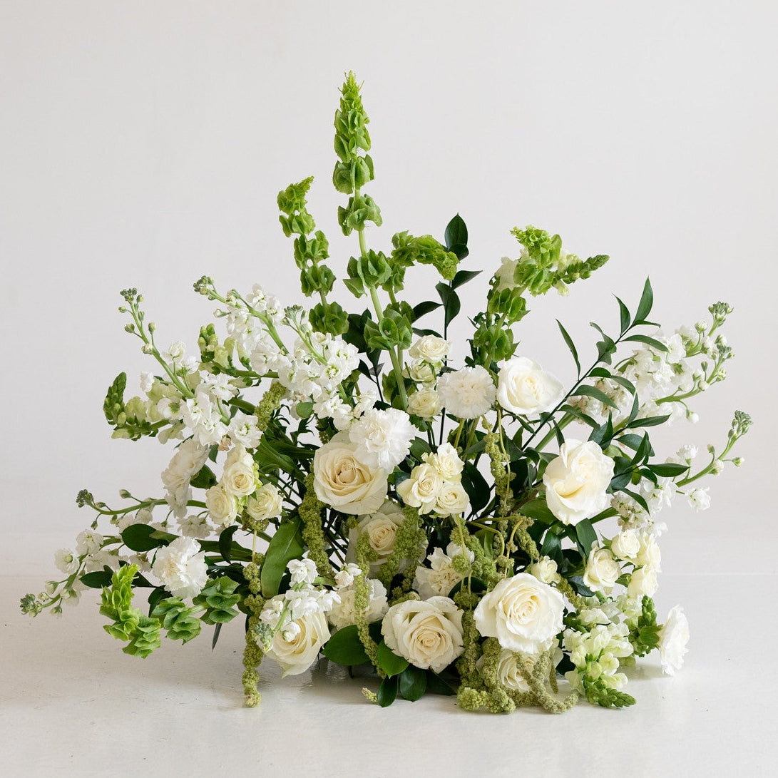 A white and green floral arrangement featuring white roses, baby's breath, and tall green foliage, photographed against a clean white studio background.