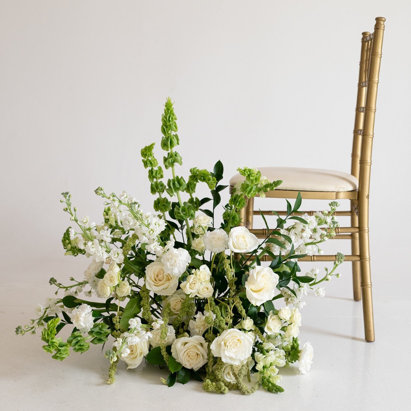 A large floral arrangement of white roses, green foliage, and white flowers placed on the floor beside a gold-framed chair against a plain off-white wall.