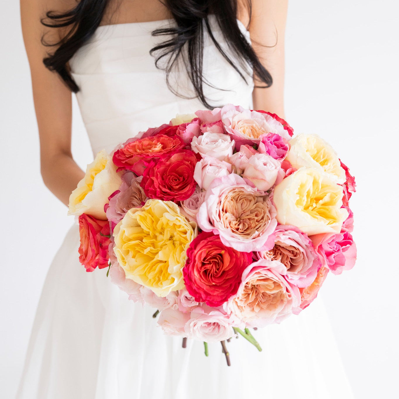A woman in a white strapless wedding dress holding a large, vibrant bouquet of multicolored roses and peonies in shades of pink, red, yellow, and peach against a clean white background.
