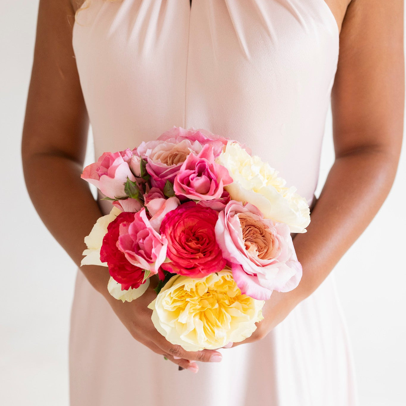 A woman wearing a pale blush pink halter-neck dress holds a vibrant bouquet of pink, red, and yellow roses against a clean white background.