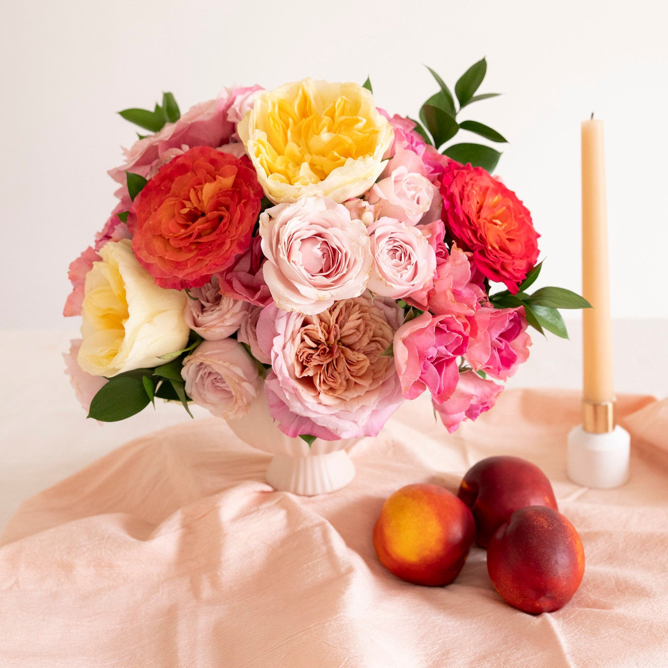 A vibrant floral arrangement of pink, red, yellow, and peach roses with green leaves, displayed in a white vase on a draped peach fabric surface next to a lit candle and three peaches.
