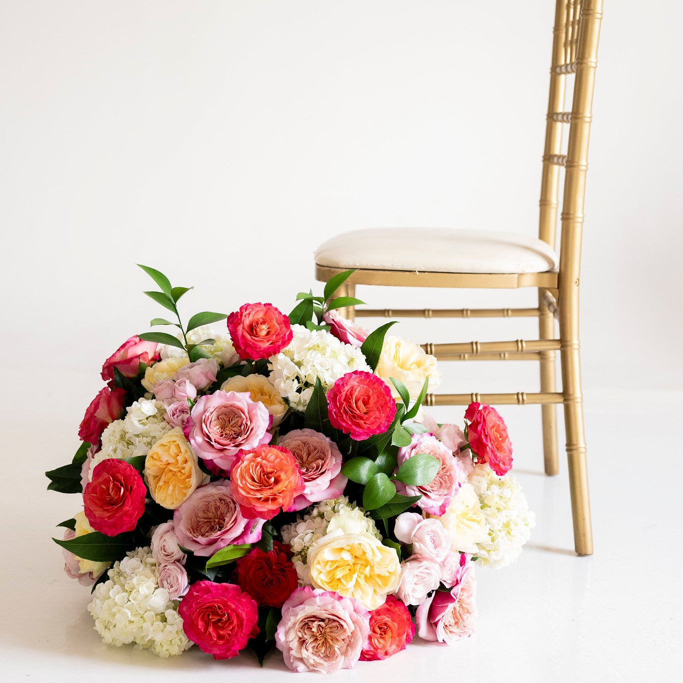 A vibrant floral arrangement of pink, red, peach, and white roses with green foliage, styled next to a gold Chiavari chair on a clean white background.