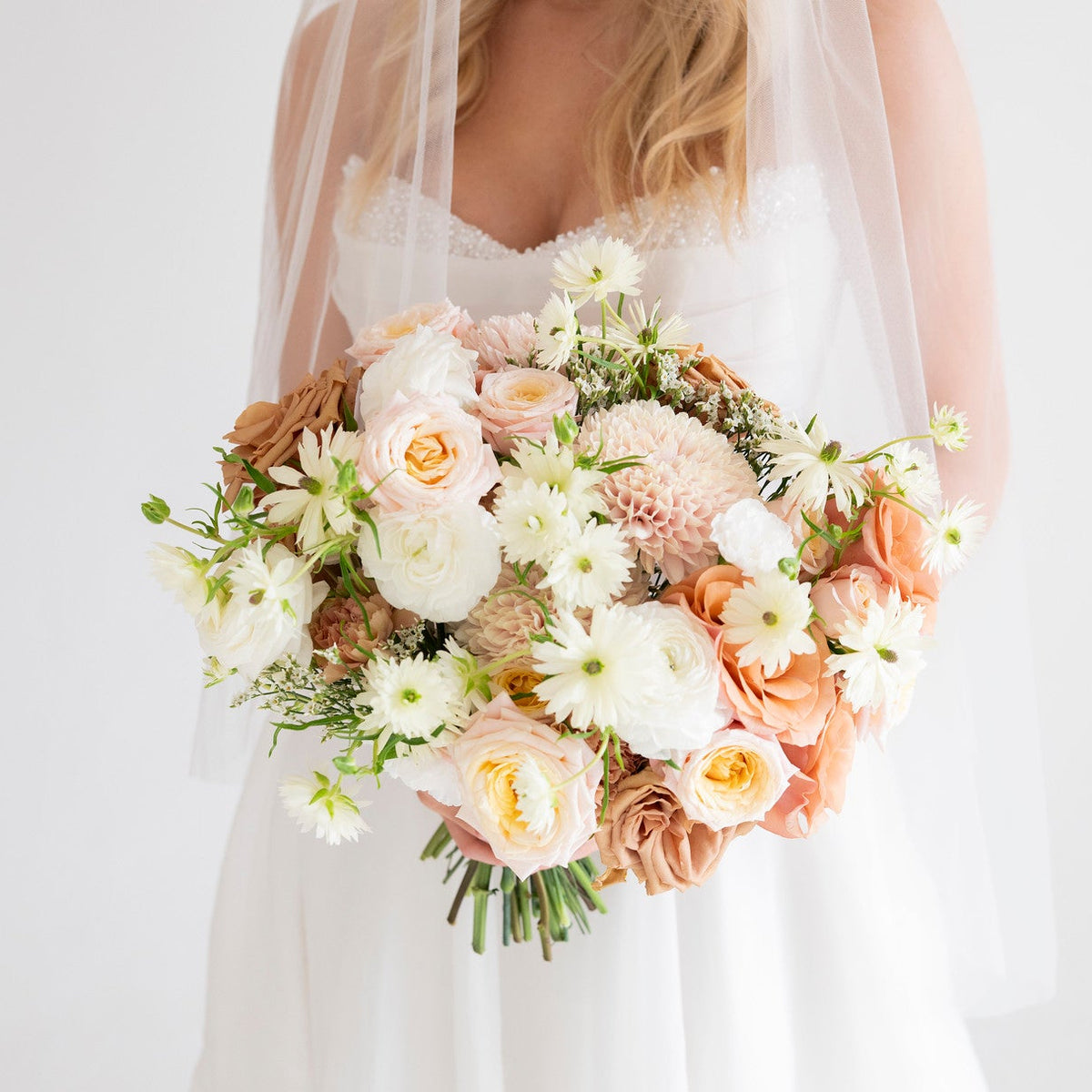 A bride in a white wedding dress and veil holding a large bridal bouquet of peach, cream, and white flowers, photographed against a soft white background.