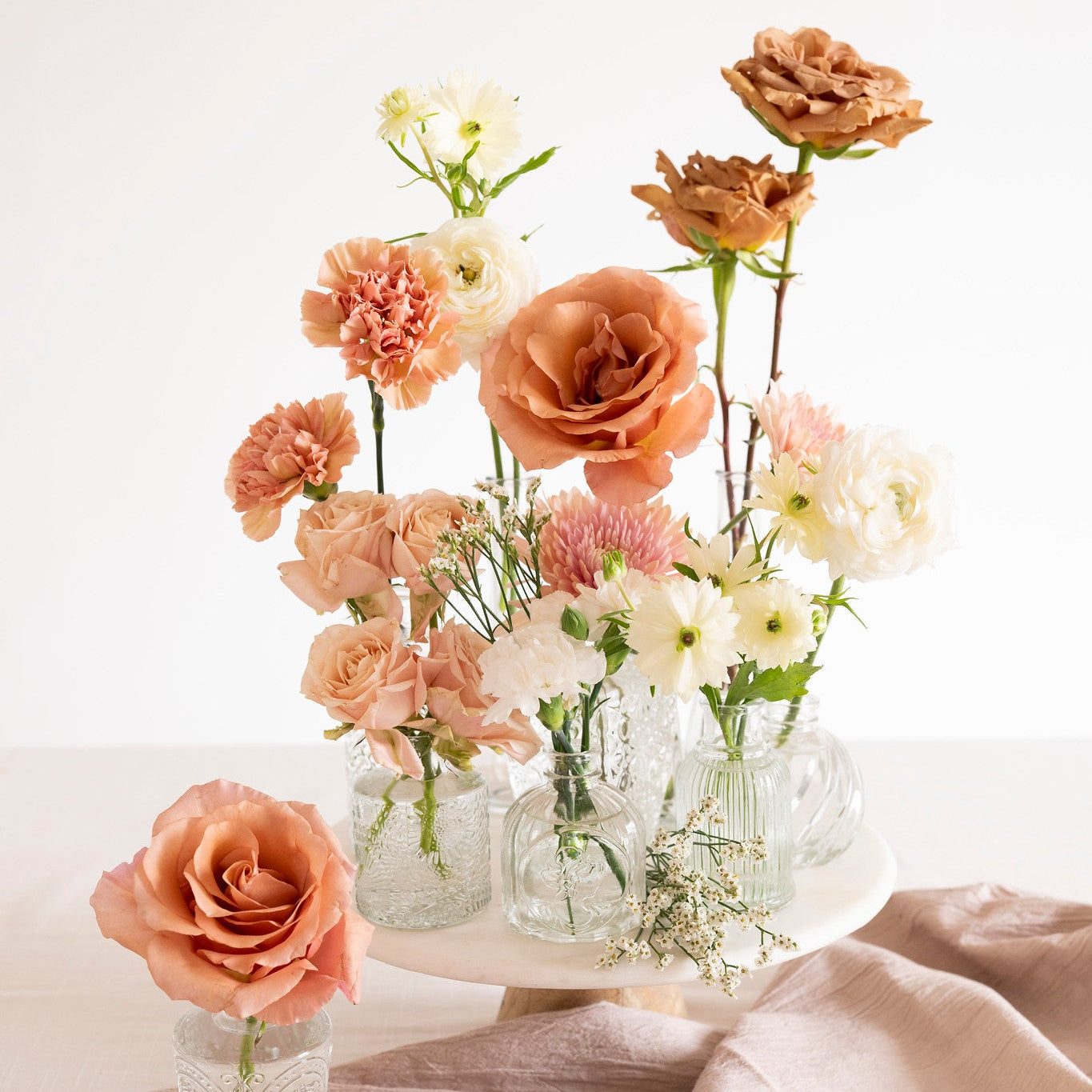 A styled floral arrangement of peach, terracotta, white, and tan flowers in clear glass vases on a white pedestal stand, with a matching peach rose in a small glass vase on a draped blush linen cloth against a white background.