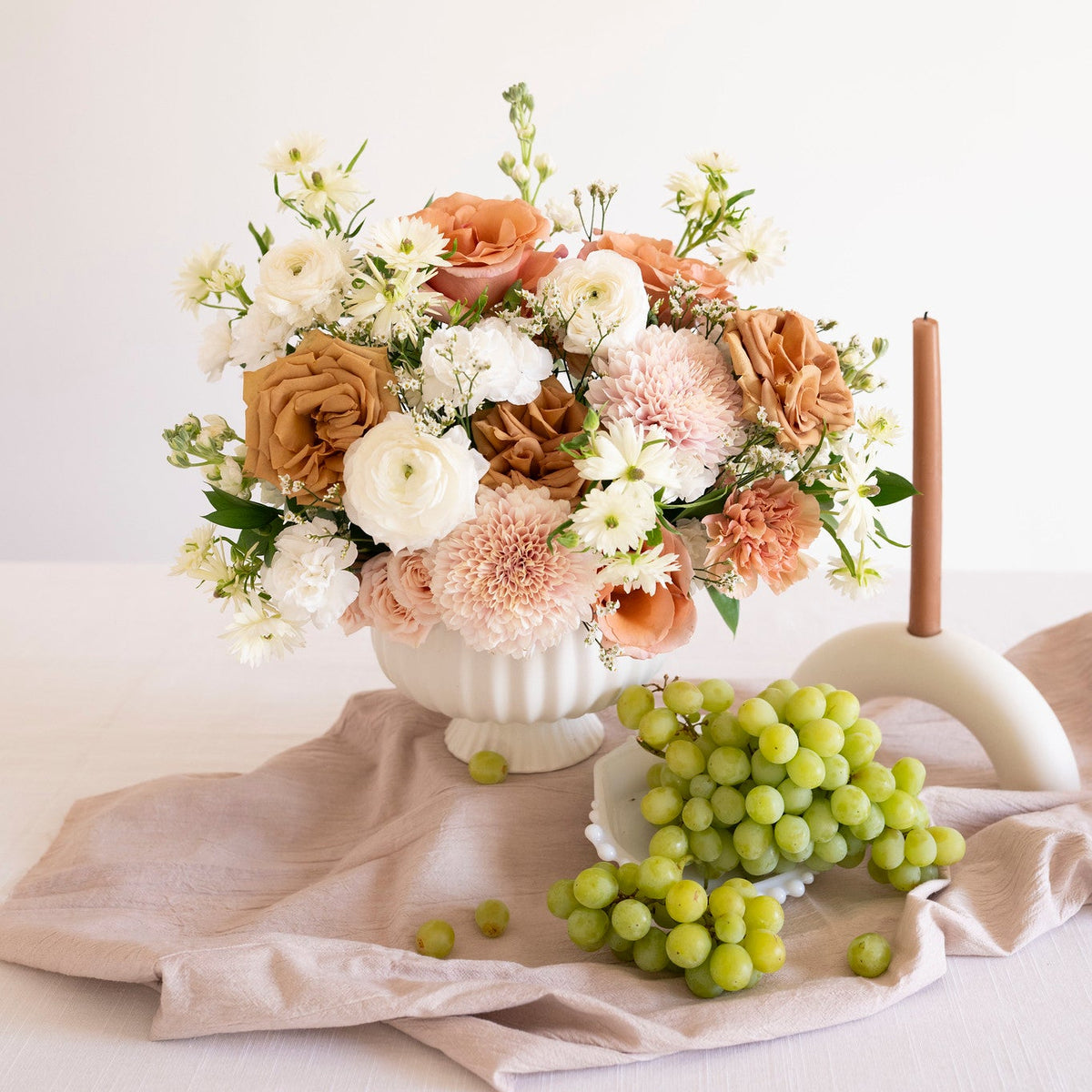 A floral arrangement in a white pedestal vase on a draped blush fabric surface, styled with green grapes and a white candle holder against a soft cream background.