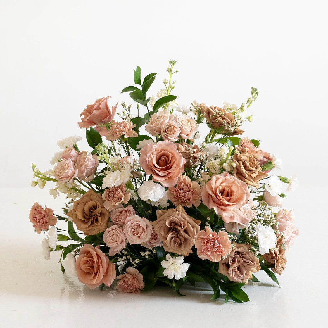 A fresh floral bouquet featuring dusty terracotta and peach roses, white filler flowers, and green foliage, arranged on a clean white surface against a white background.