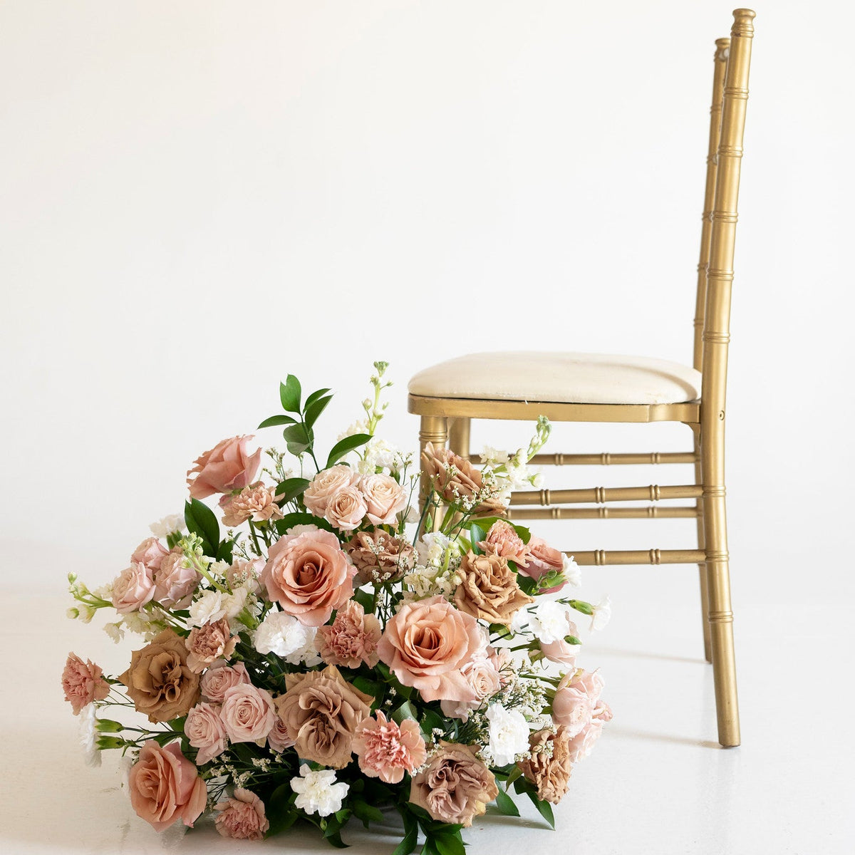 A large floral arrangement in blush pink, peach, and white with green foliage sits beside a gold bamboo-style chair against a clean off-white studio background.
