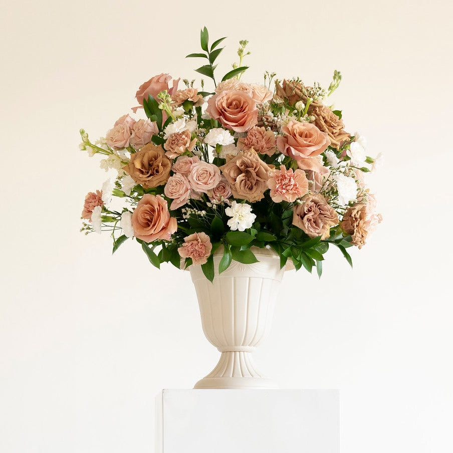 A bouquet of terracotta roses and green foliage arranged in a white urn-shaped vase, displayed on a tall white pedestal against a clean white background.