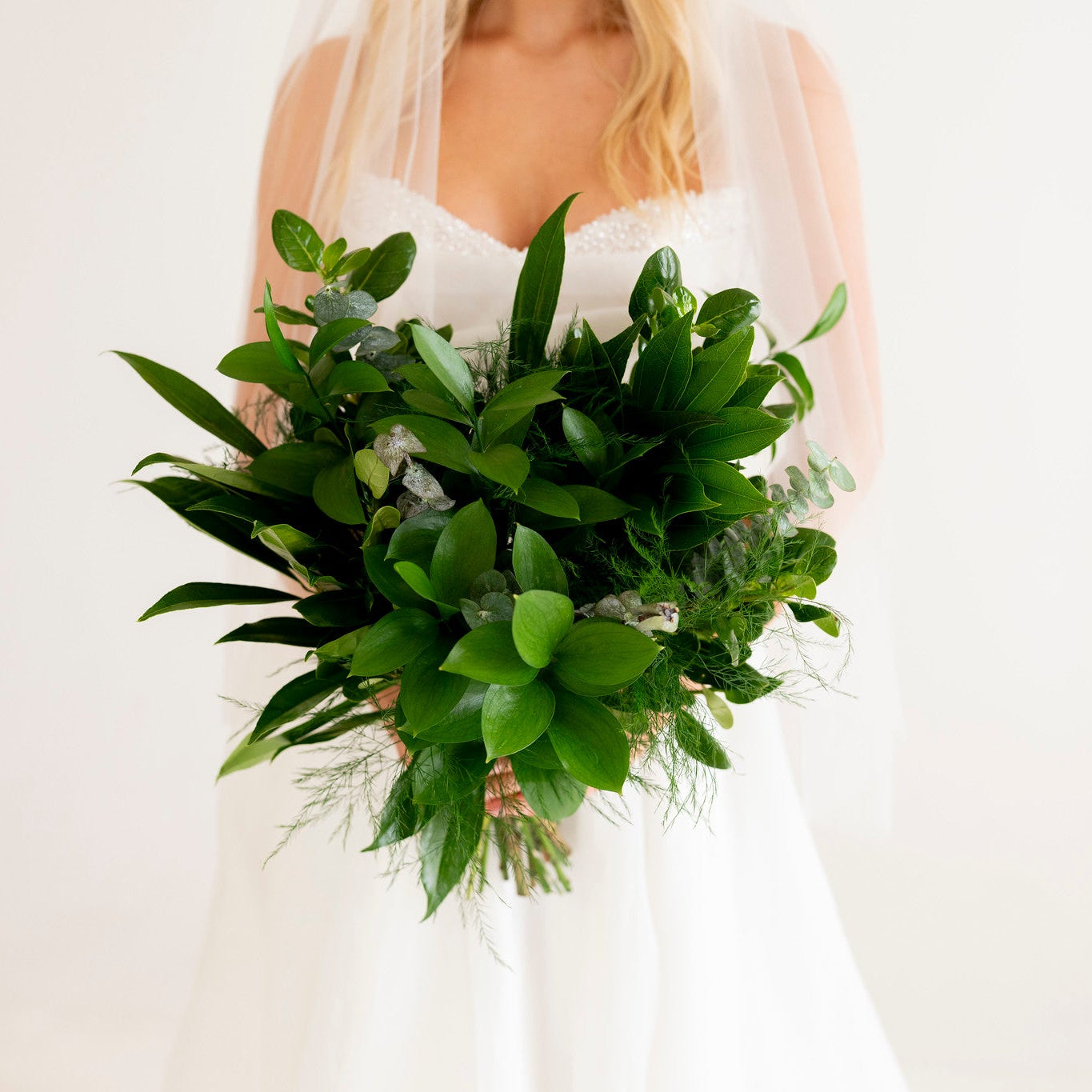 A bride in a white wedding dress and veil holding a lush green floral bouquet against a soft white background.