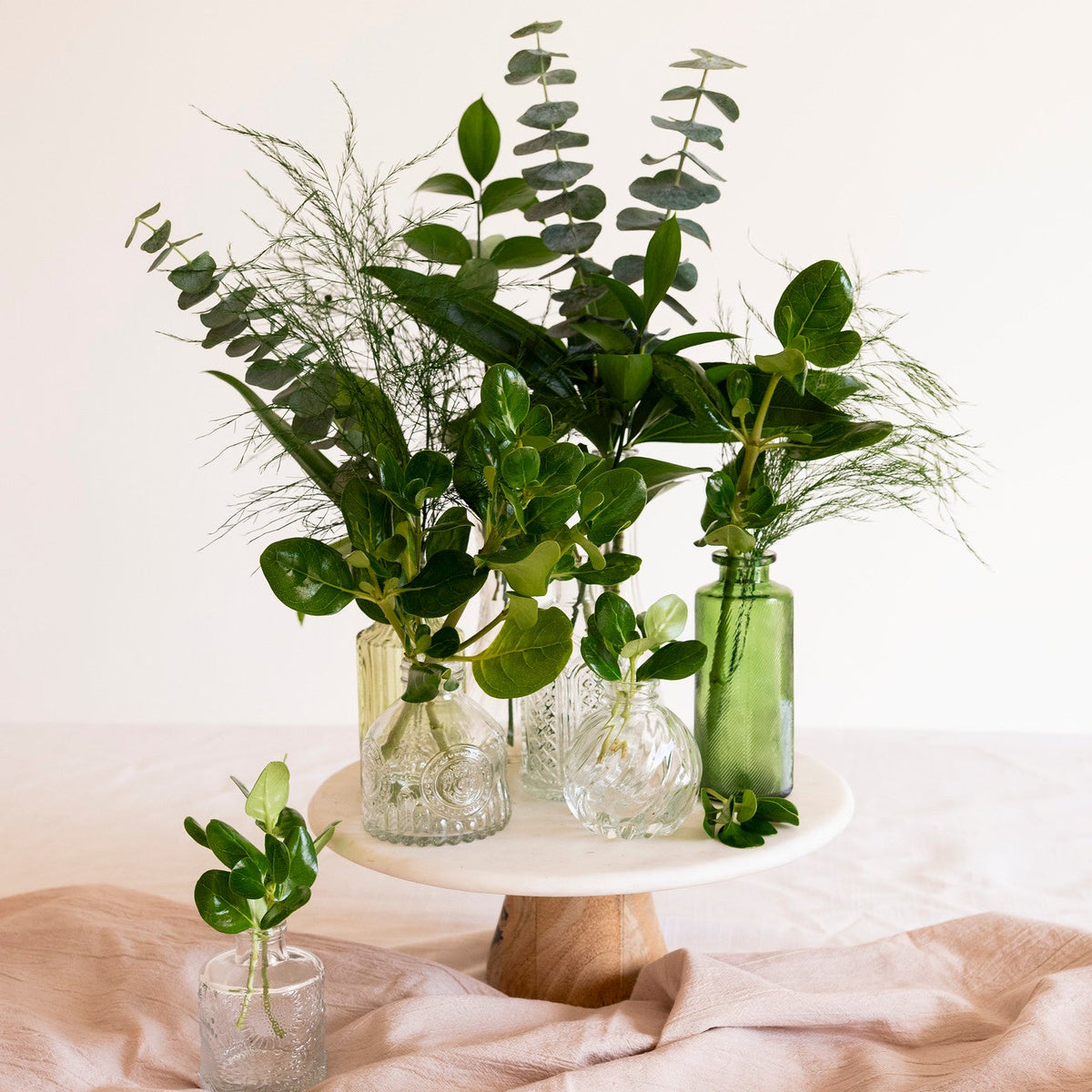 A styled floral arrangement of green eucalyptus and other greenery in clear glass bottles and a small clear glass cup, displayed on a white pedestal cake stand with a blush pink fabric drape against a warm off-white background.